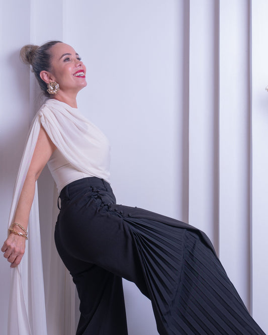 A woman posing in a white draped top and black pleated wide-leg trousers against a white architectural background with a vase of flowers.